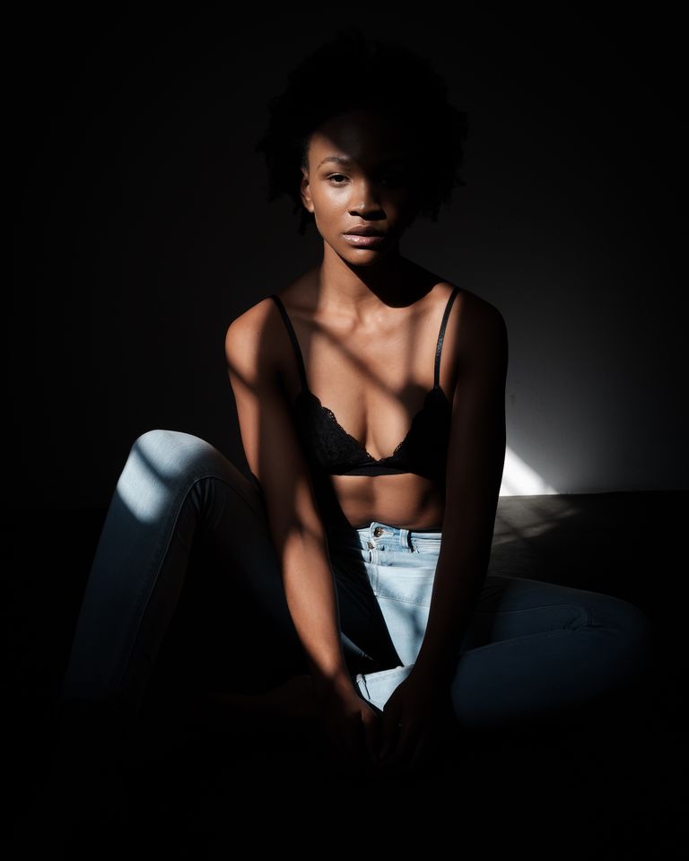 A black woman sitting in the dark and wearing a bra with jeans 